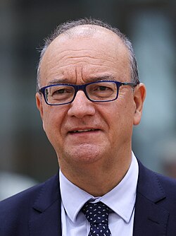 Head-and-shoulders portrait of Giuseppe Valditara in 2025, wearing dark-rimmed glasses, a navy suit and patterned tie