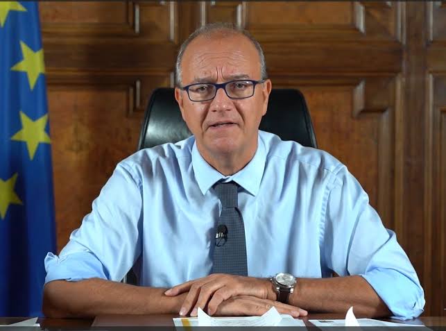 Giuseppe Valditara seated at his desk recording a video address, wearing a light-blue shirt and tie, with the flag of the European Union behind him
