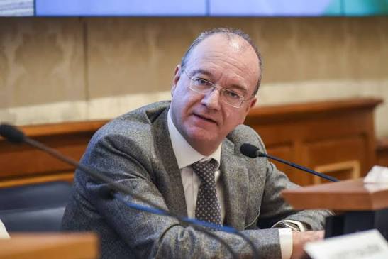 Giuseppe Valditara seated at a conference table with a microphone, in a grey jacket