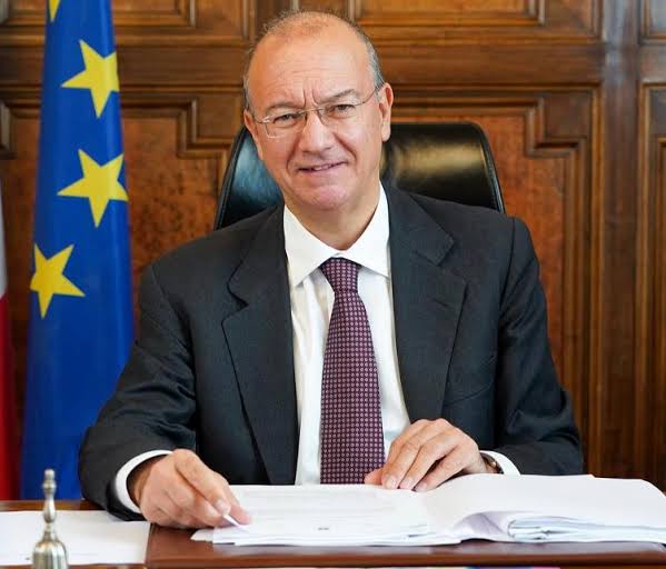 Giuseppe Valditara seated at a desk with papers and a small bell, smiling, in a dark suit and burgundy tie