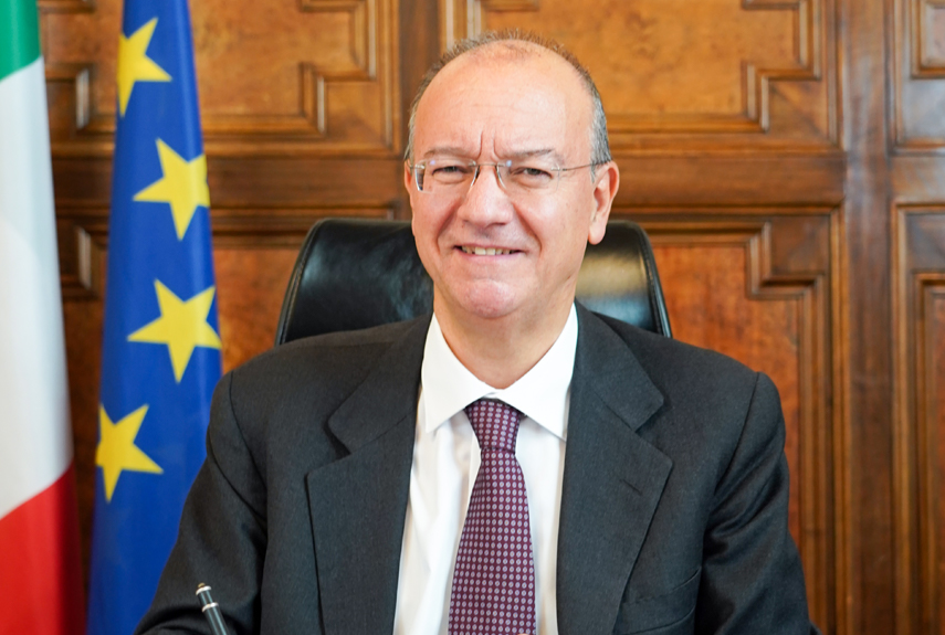 Giuseppe Valditara smiling at his desk in a wood-panelled ministerial office, with the Italian and European flags behind him