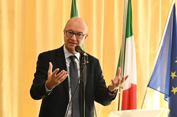 Giuseppe Valditara speaking at a microphone with open hands, Italian and European flags behind him against a warm yellow curtain