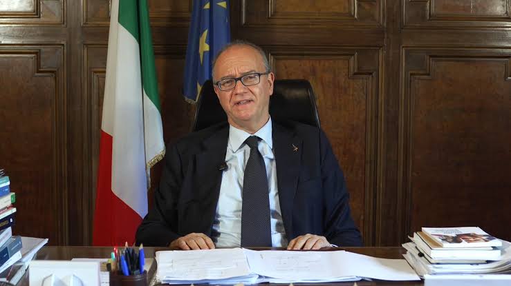 Giuseppe Valditara seated at a large desk with papers and books in a wood-panelled office, the Italian and European Union flags behind him