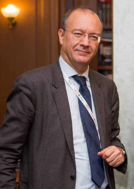 Giuseppe Valditara in a grey suit and striped tie, wearing a lanyard, standing in a room lined with bookshelves