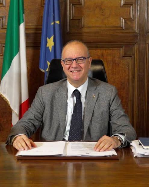 Giuseppe Valditara in a grey suit, smiling at his desk with open documents in front of him and flags behind