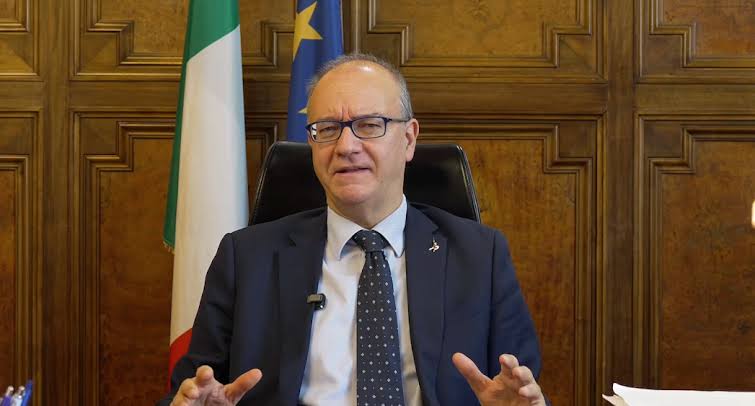 Giuseppe Valditara seated at a desk, gesturing with his hands as he speaks, with flags behind him