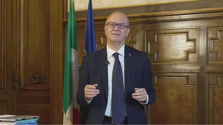 Giuseppe Valditara standing in a wood-panelled office, gesturing with one hand as he addresses the camera, with flags behind him