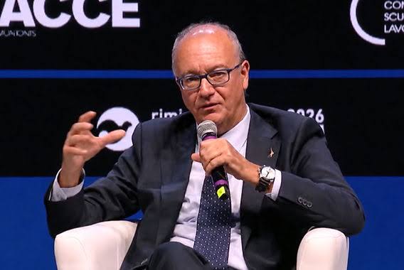 Giuseppe Valditara seated in a white armchair on a stage with a blue backdrop, speaking into a handheld microphone at the Meeting di Rimini