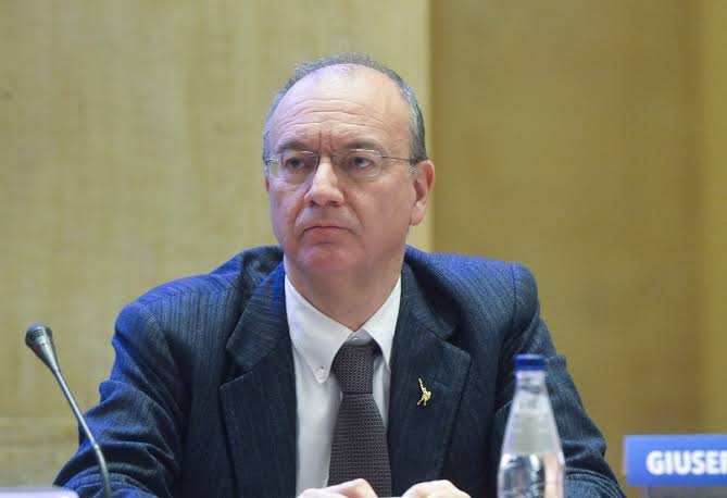 Giuseppe Valditara seated at a conference panel table, speaking into a microphone, with a nameplate partly visible in front of him