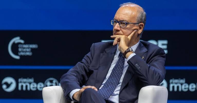Giuseppe Valditara seated in a white armchair on stage at the Meeting di Rimini, hand to his chin, listening attentively to a speaker
